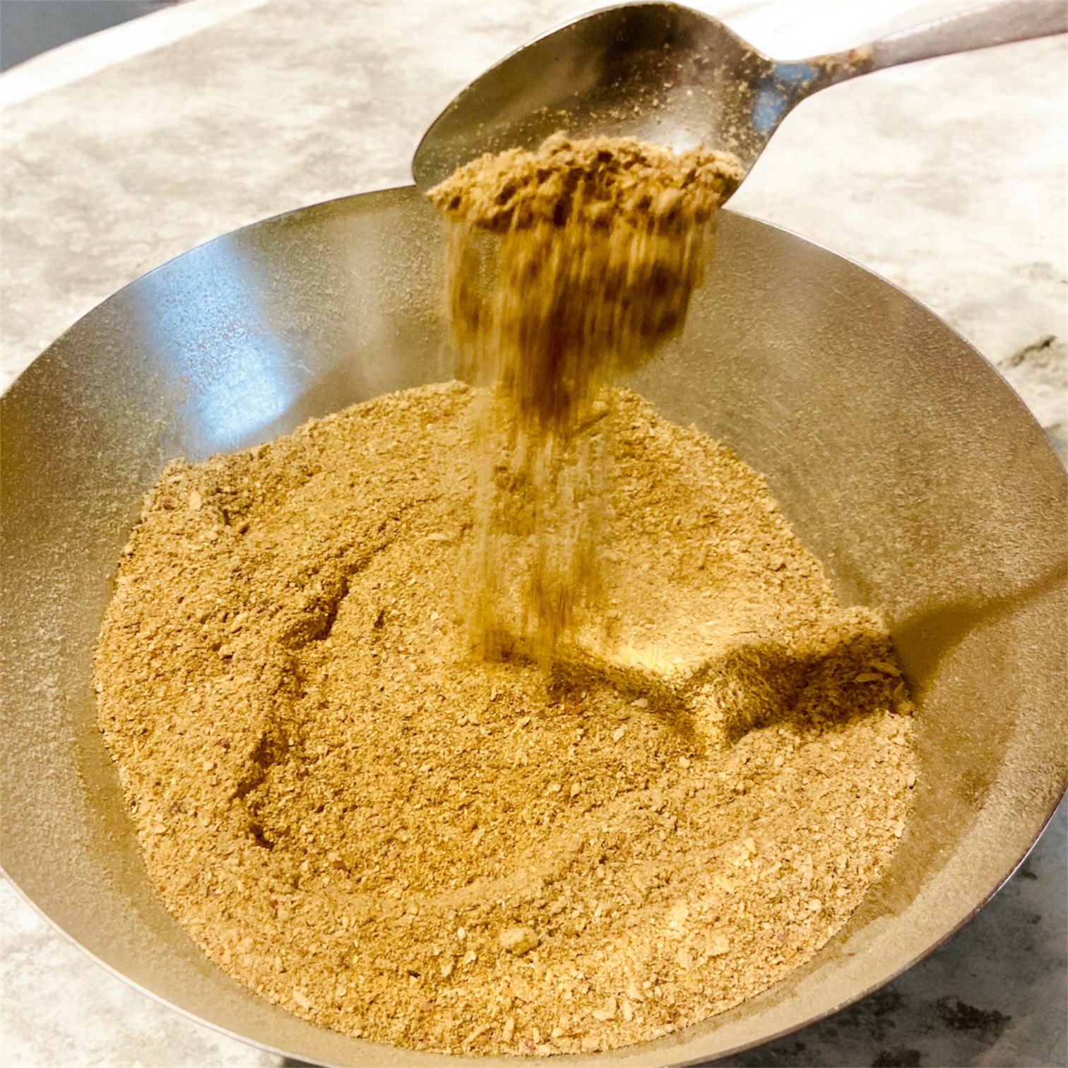 Un saladier en métal posé sur un plan de travail clair, rempli d’une poudre d’assaisonnement aux champignons, de couleur beige doré. Une cuillère au-dessus du saladier laisse couler la poudre en un fin filet, créant un léger relief à la surface. La texture est fine et homogène, évoquant un condiment sec prêt à être utilisé en cuisine.